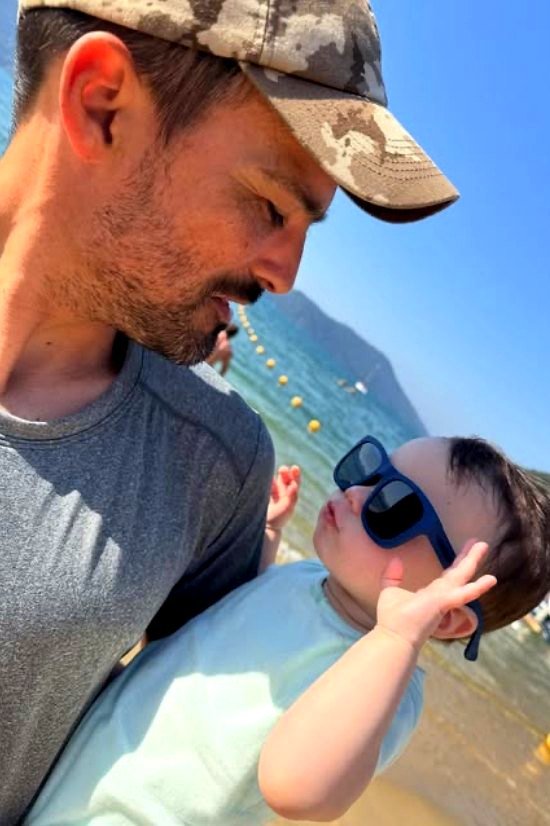 Leo Nardi Borba à beira-mar, de boné, olhando para o filho de óculos escuros