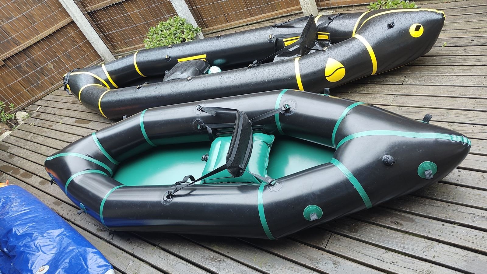 Dois packrafts inflados sobre um deck de madeira: um preto com faixas verdes e fundo verde, outro maior preto com faixas amarelas